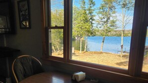 A dining area with a round table and chairs, a window with a view of a lake and trees, and a framed picture on the wall. - Cabin with private peninsula on Presque Isle Chain of Lakes! (Presque Isle)