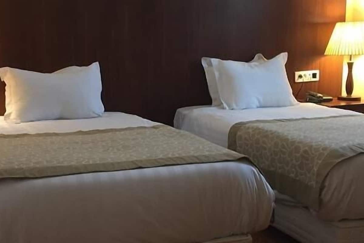 A hotel room with two beds, each with a pillow and a blanket. There are bedside lamps on either side of the beds.