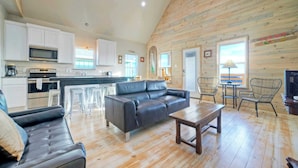 A spacious living area with a black leather sofa, wooden coffee table, and wicker chairs. A kitchen with white cabinets and a microwave is visible in the background. - 2 Homes, Sleeps 16, Shared Pool+boat Dock Laguna Village Serenity by Avantstay (Corpus Christi)