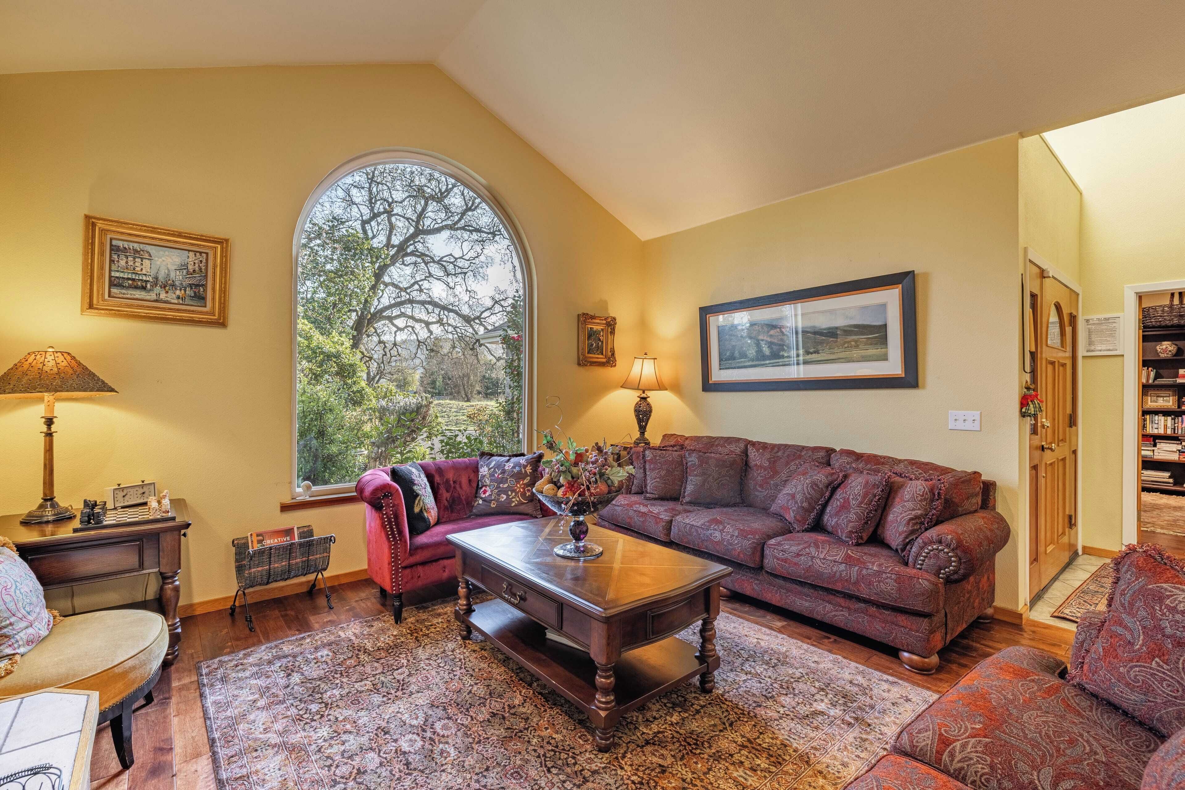 A cozy living room with a large window, a red sofa, a wooden coffee table, and a bookshelf.