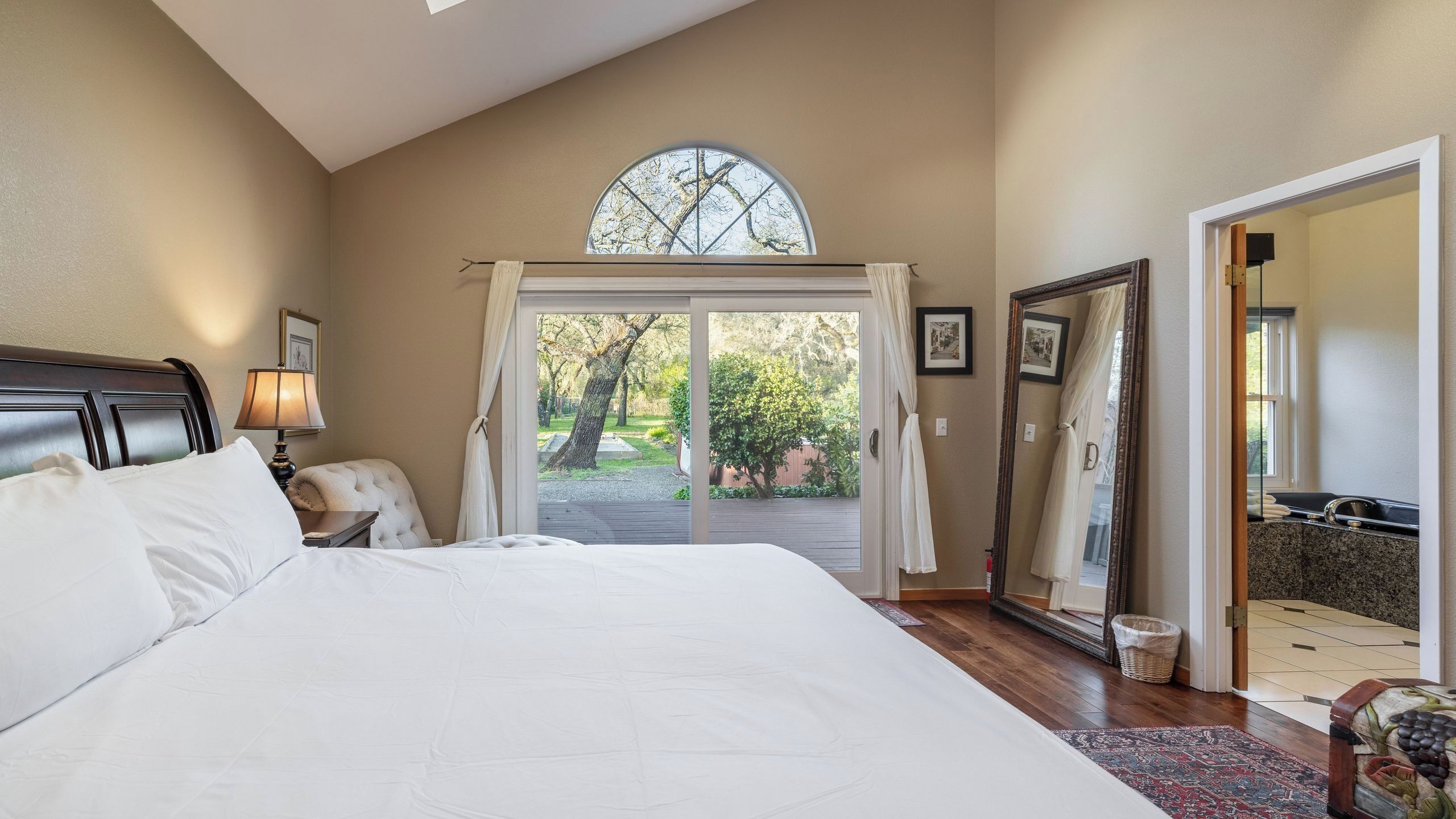 A bedroom with a large bed, a nightstand, a lamp, a window with curtains, a door leading to a bathroom, and a view of the outdoors.