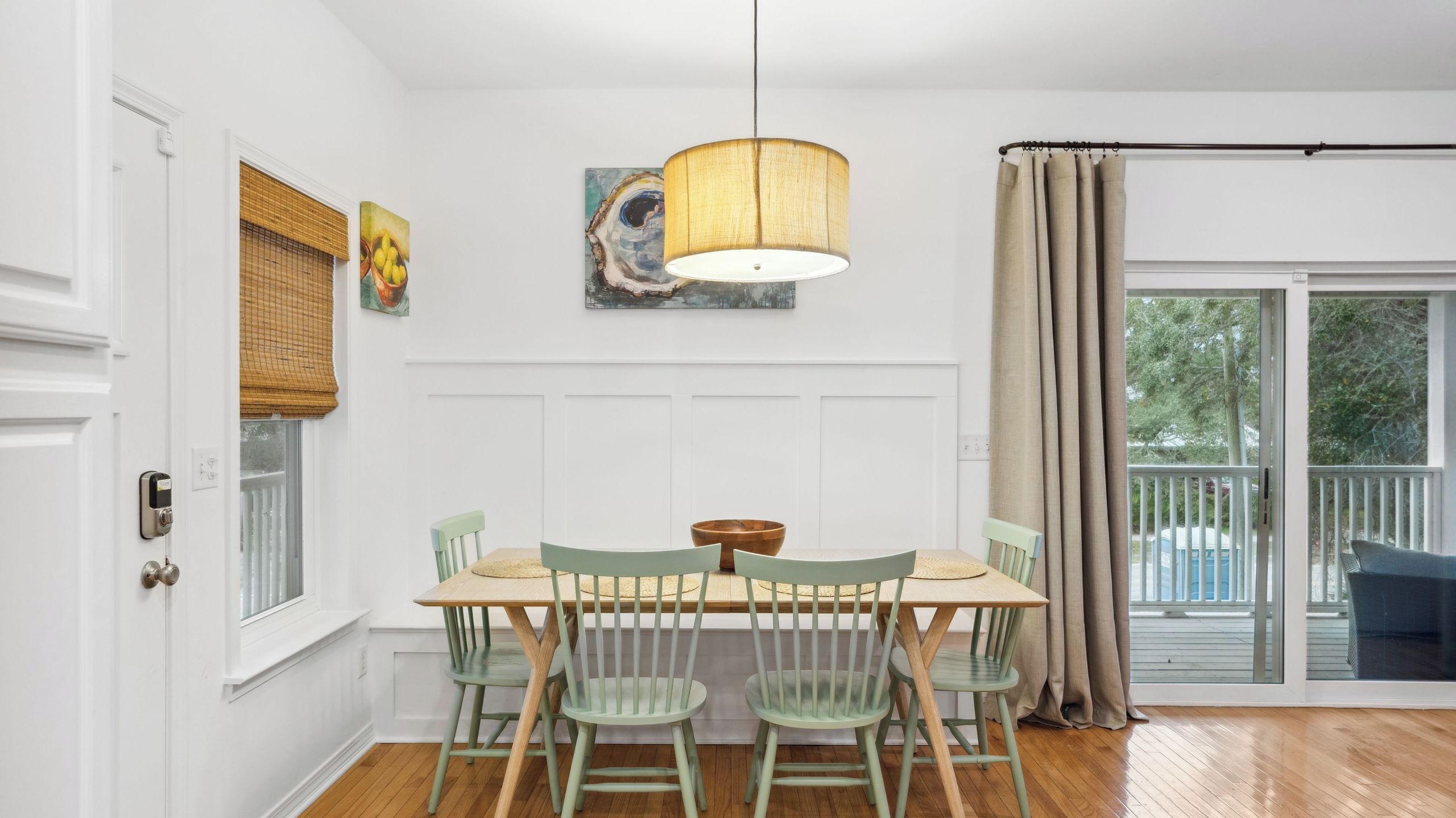Un coin repas avec une table en bois, quatre chaises, un luminaire suspendu et une vue sur un balcon à travers une porte vitrée.