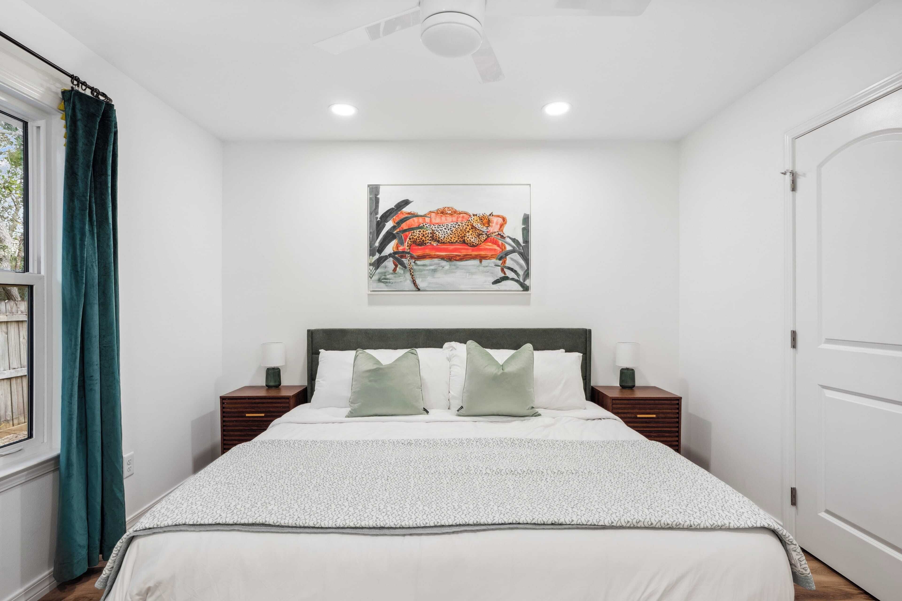 A bedroom with a bed, a headboard, two bedside tables, a nightstand, a ceiling fan, a window with curtains, and a painting on the wall.