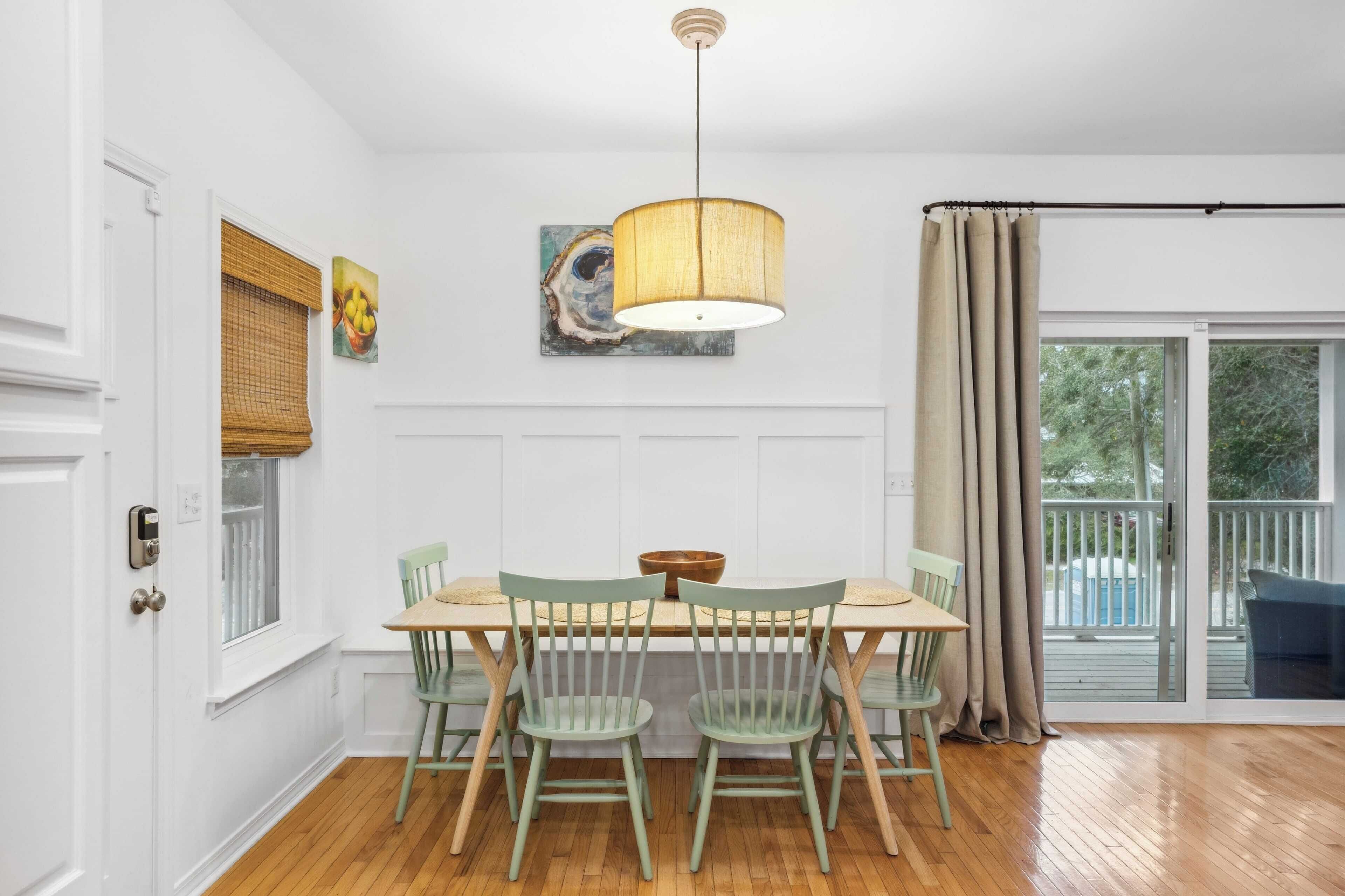 Un coin repas avec une table en bois et quatre chaises, une lampe suspendue et une fenêtre avec des stores.