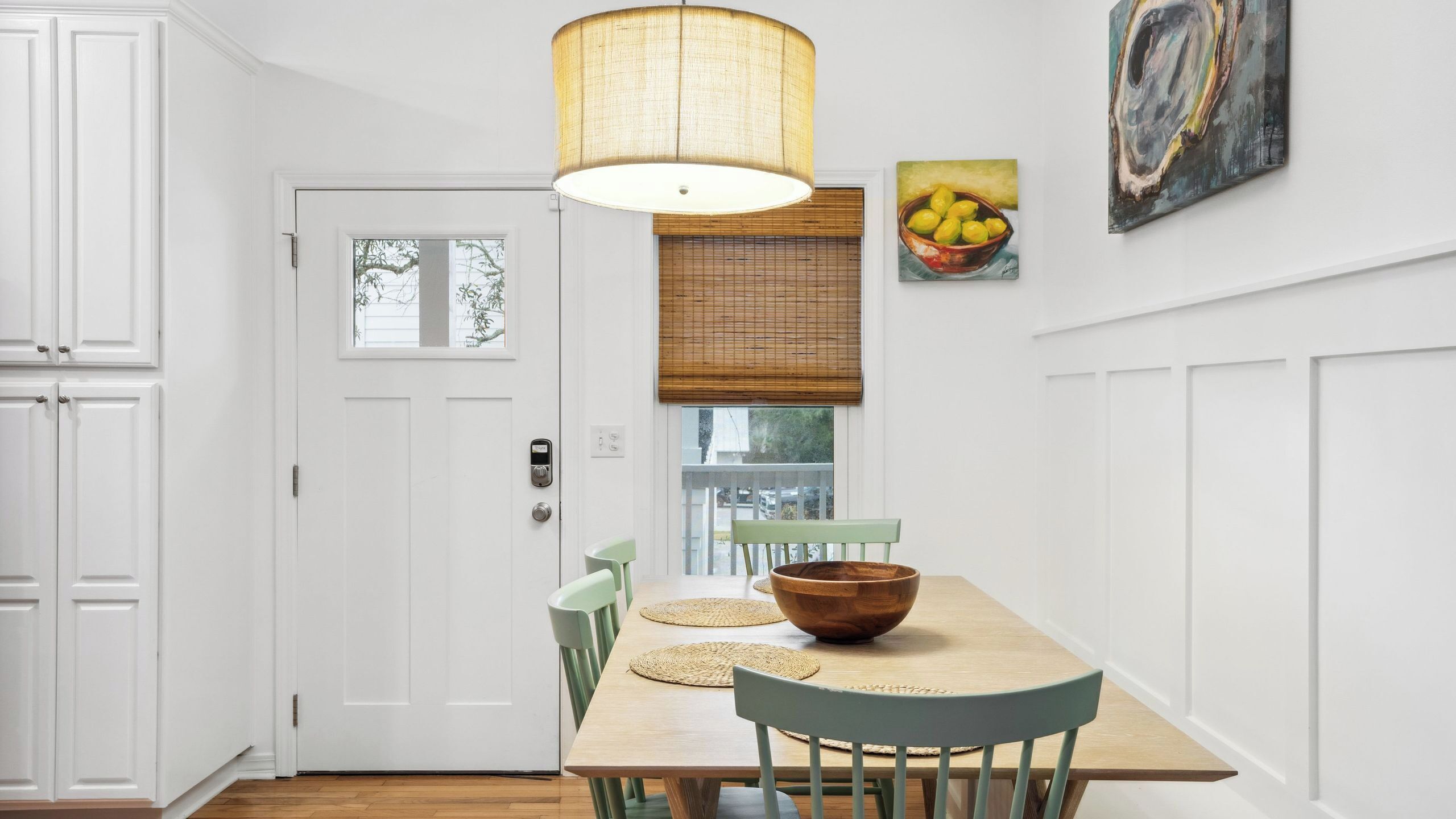 A dining area with a wooden table, chairs, a bowl, a hanging lamp, and a window with blinds.