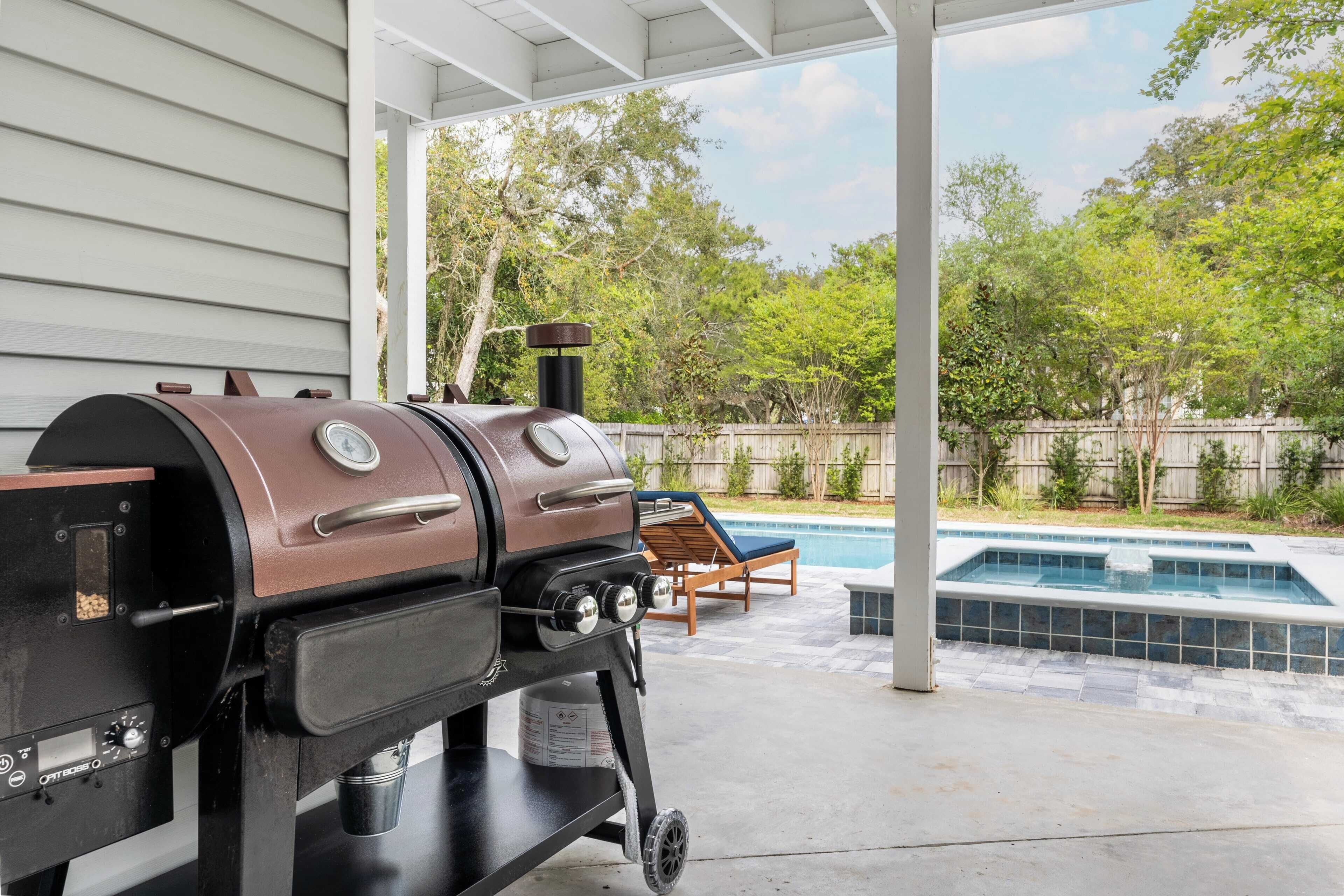 A barbecue grill with a digital thermometer and a water tank, positioned under a covered patio area with a pool and lounge chairs in the background.
