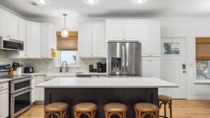 A modern kitchen with white cabinetry, stainless steel appliances, a large island with stools, and a window with a view. - Dolphin Days by Avantstay Pool, 0.2mi to Beach! (Destin)