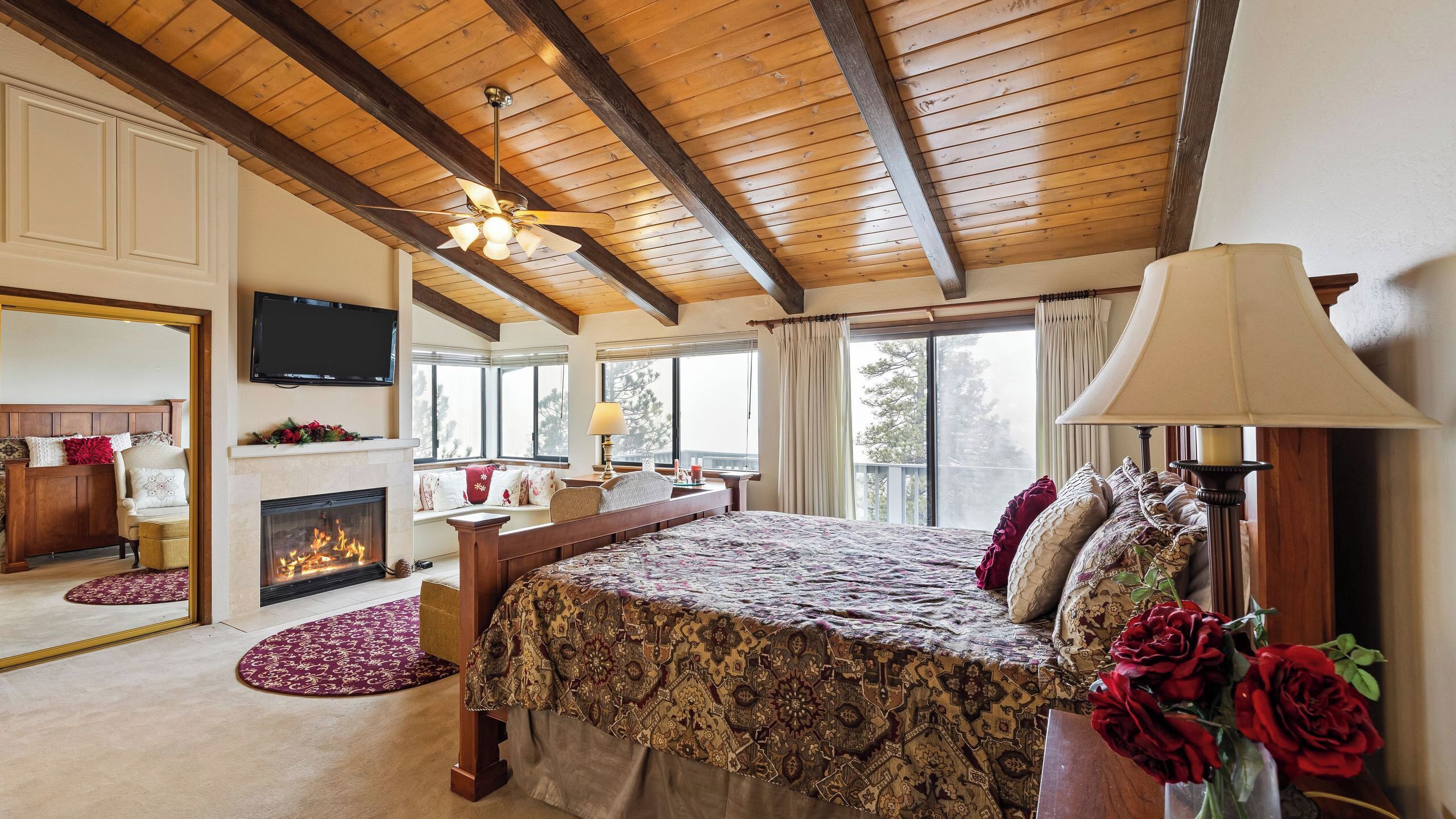 A bedroom with a fireplace, a bed with patterned bedding, a desk with a lamp, and a television on the wall.