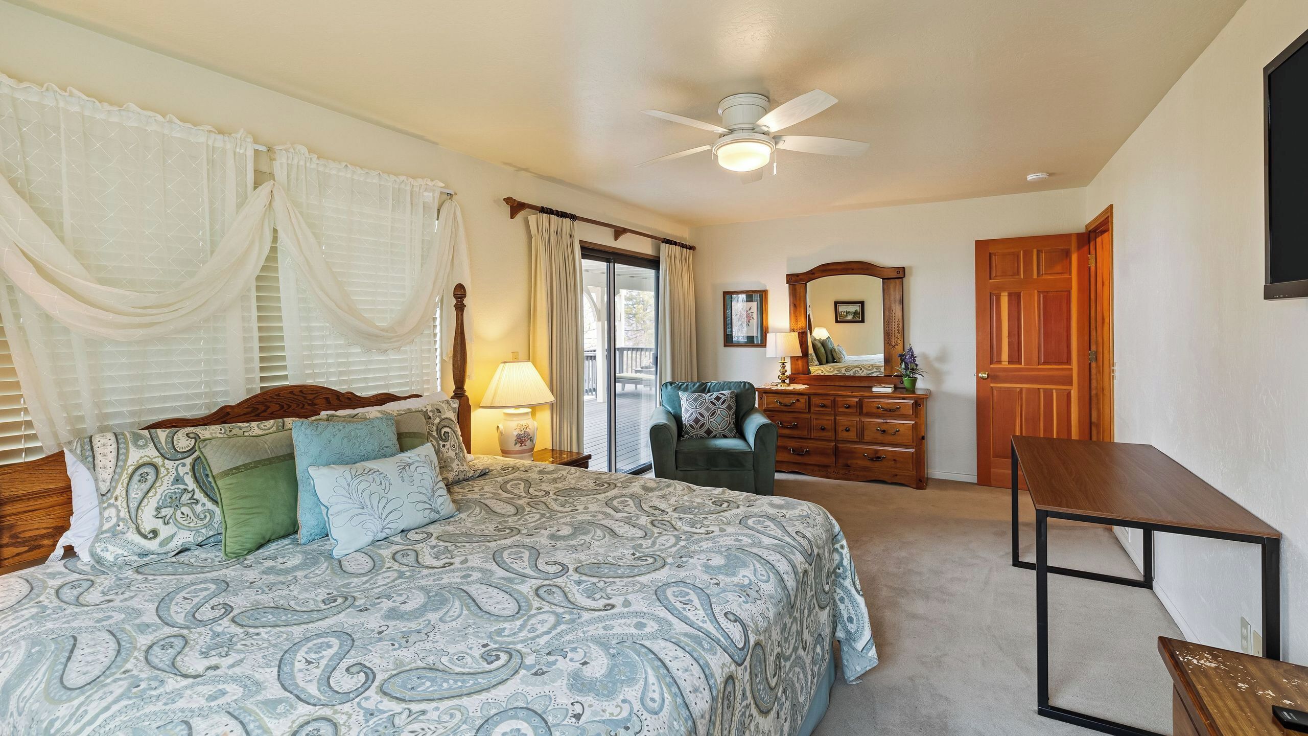 Une chambre à coucher comprenant un lit, une chaise, une commode, un bureau, un ventilateur de plafond et une porte.