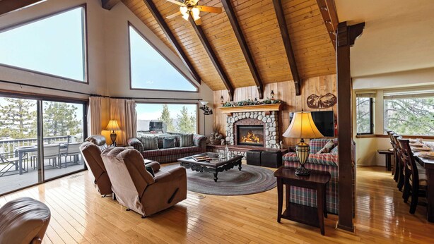 A spacious living room with a fireplace, wooden ceiling beams, and large windows offering a view of the outdoors. - 6bdr Group Retreat, Decks w/ Incredible Views Starry Heights by Avantstay (Lake Arrowhead)