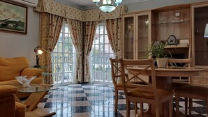 A living room with a checkered floor, a sofa, a glass coffee table, and a wooden dining table with chairs. There are curtains, a painting, and a cabinet with glass doors.