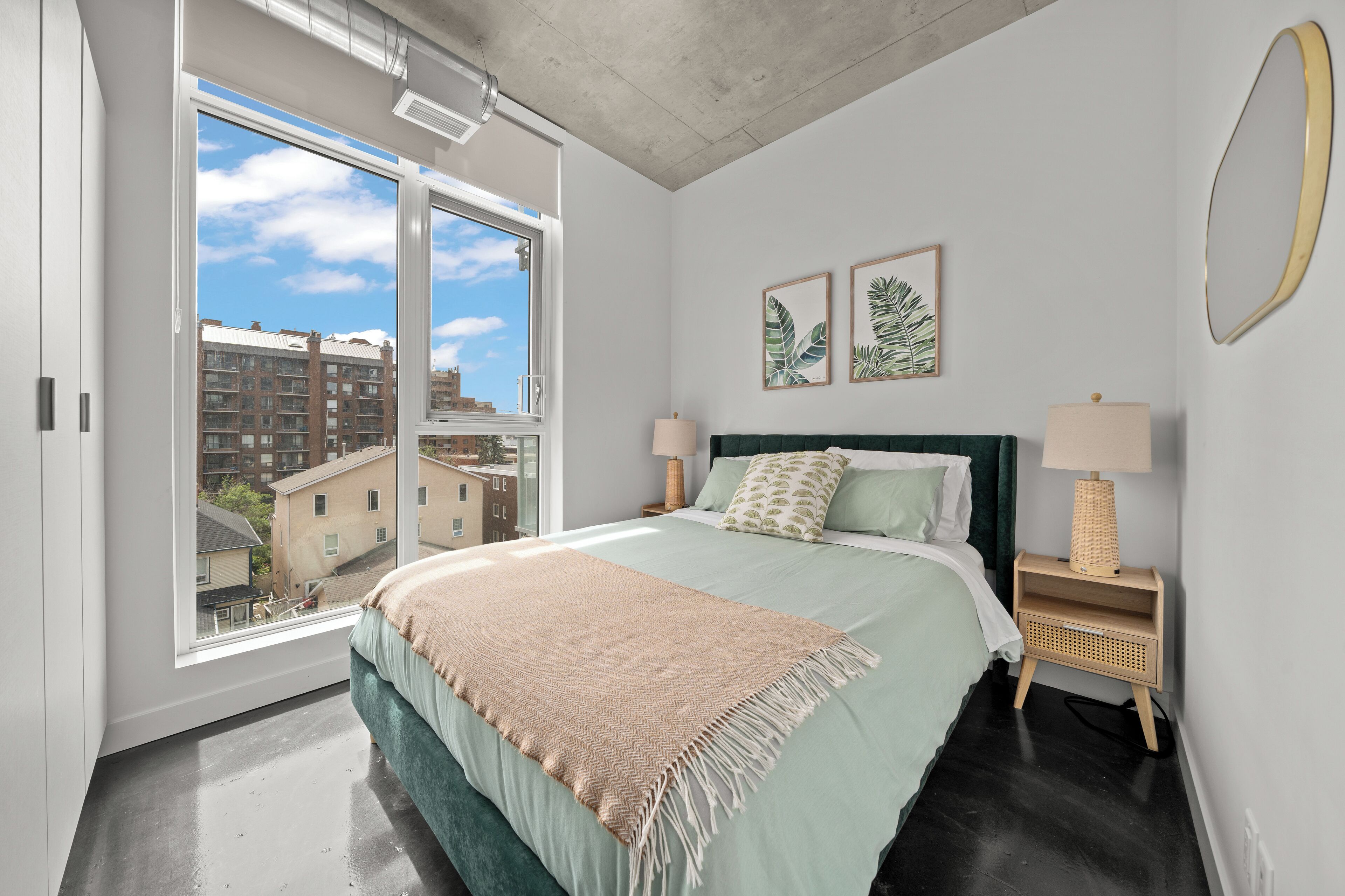 A bedroom with a large window, a bed with a green headboard, two bedside tables with lamps, and two framed leaf prints on the wall.