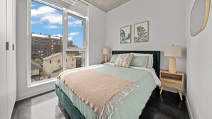 A bedroom with a large window, a bed with a green headboard, two bedside tables with lamps, and two framed leaf prints on the wall. - Cosmopolitan Calgary, Modern Loft (Calgary)