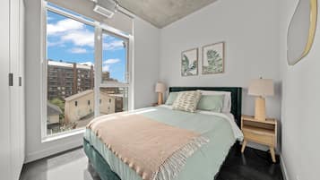 A bedroom with a large window, a bed with a green headboard, two bedside tables with lamps, and two framed leaf prints on the wall.