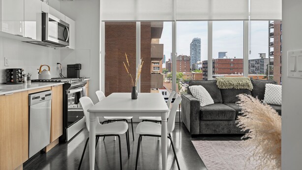 A modern kitchen with white appliances, a dining area with a white table and chairs, and a grey sofa with a view of the city through large windows. - Cosmopolitan Calgary, Modern Loft (Calgary)