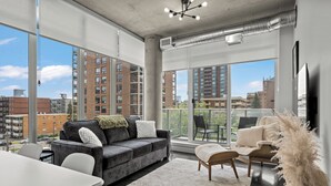 A modern living room with a grey sofa, white armchair, and a large window offering a city view. - Cosmopolitan Calgary, Modern Loft (Calgary)