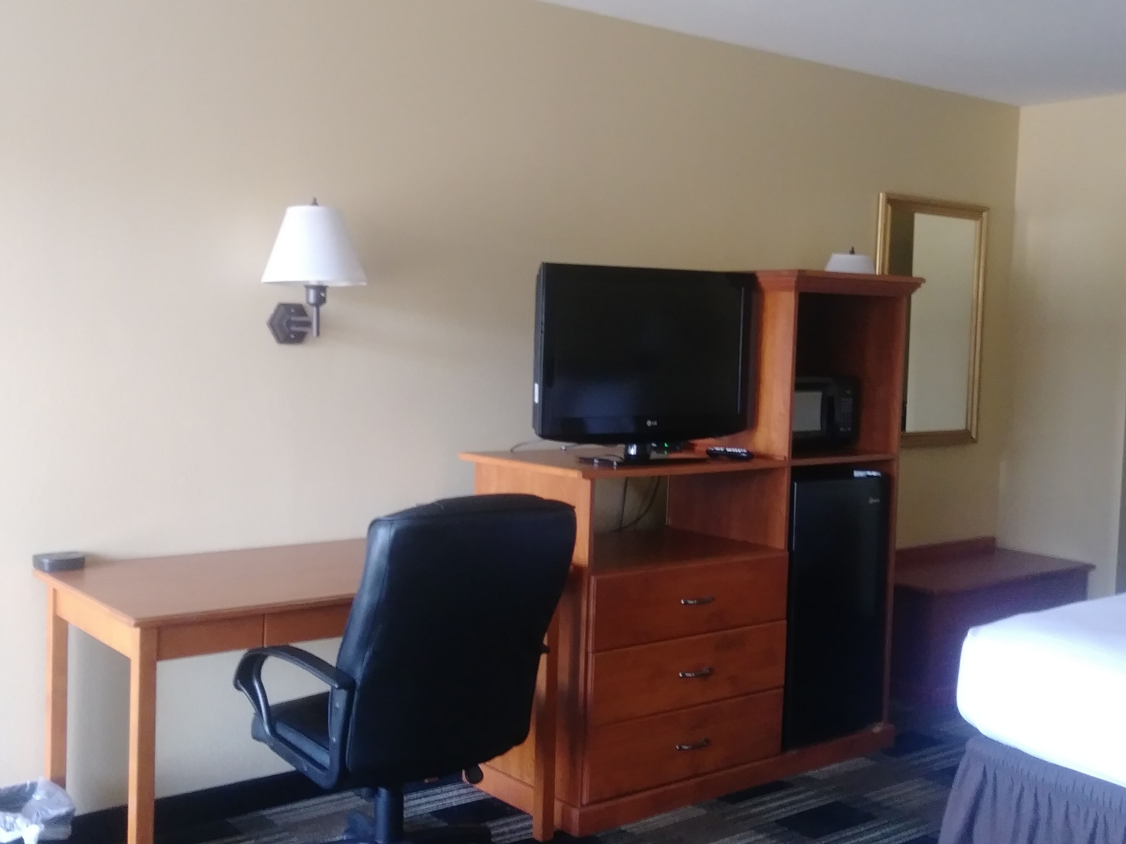 A hotel room with a wooden desk, a black office chair, a television on a wooden stand, a small cabinet, a lamp, and a bed with a white cover.