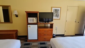 A hotel room with two beds, a wooden dresser, a microwave, a television, a small refrigerator, a lamp, and a framed picture on the wall.