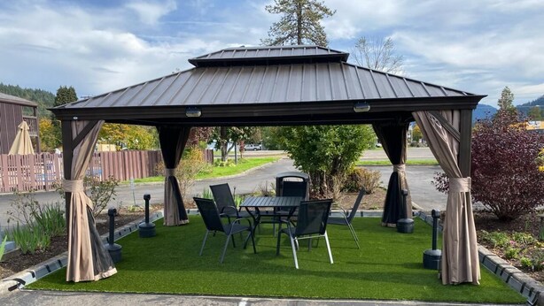 A gazebo with a metal roof and four chairs, surrounded by a small patch of grass.