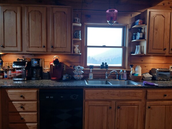 A kitchen with wooden cabinets, a coffee maker, a kettle, a sink, and a window with a view of trees.