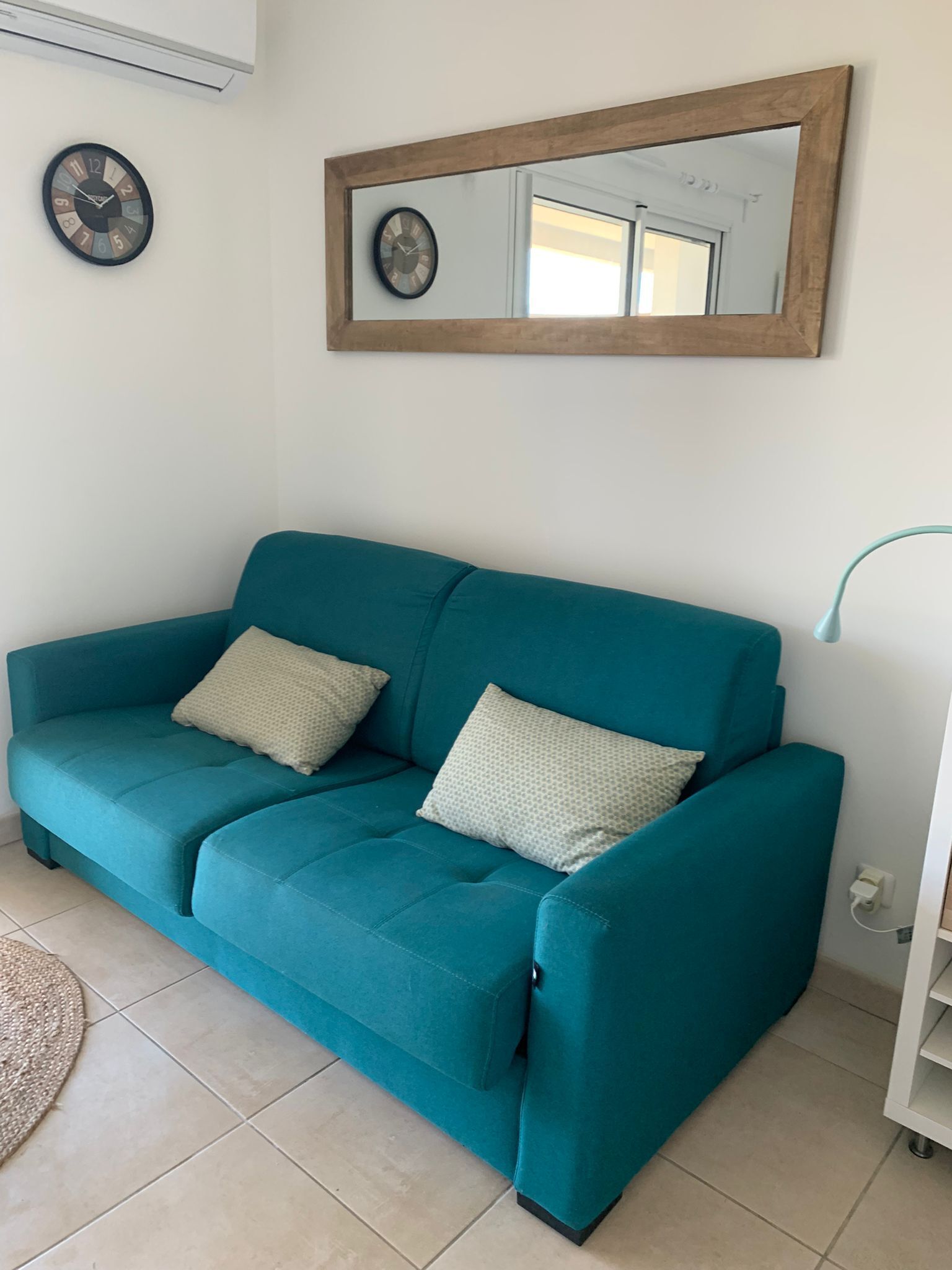 Ein blaugrünes Sofa mit zwei Kissen, einem spiegelnden Holzrahmen und einer an der Wand befestigten Uhr.