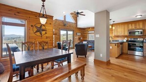 A wooden interior with a dining area, a black leather chair, a kitchen with modern appliances, and a view through large windows.