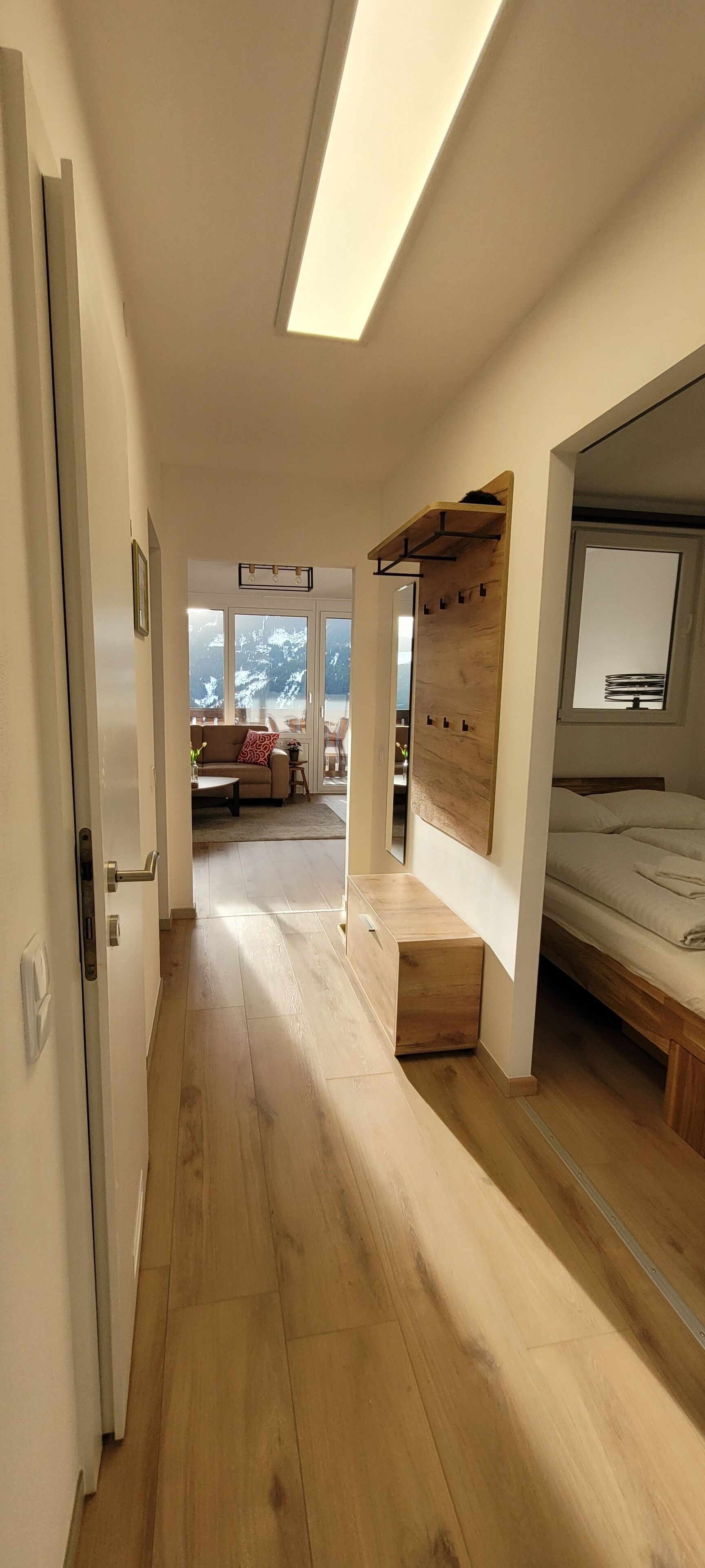 Um interior moderno com piso de madeira, uma cama com cabeceira, uma prateleira de madeira fixada na parede e vista para uma sala de estar com sofá e mesa.