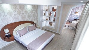 A hotel room with a large bed, a bedside table with a lamp, a phone, and a decorative shelf with pottery. - Rue d'Or (Setif)