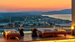 A rooftop terrace with a sunset view, two lounge chairs, a small table with a drink, and a bowl of fruit.