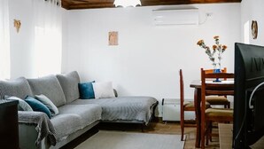 Ein Wohnzimmer mit Holzboden, einem grauen Sofa, einem Esstisch mit Stühlen und einem Fernseher auf einer Holzkommode.
