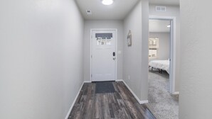 Un pasillo estrecho con una puerta blanca, piso de madera oscura y una habitación alfombrada al fondo.
