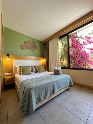 A hotel room with a bed, bedside tables, a wall decoration, a window with a view of pink flowers, and a nightstand. - Andoya Pension (Bodrum)