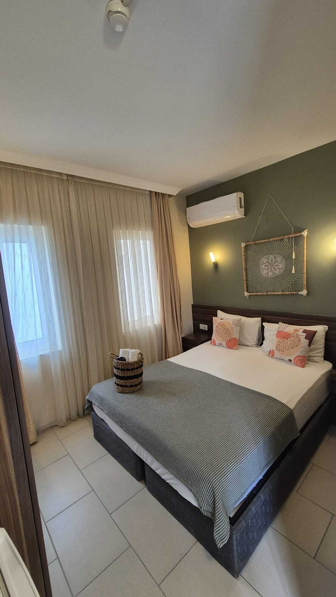 A hotel room with a large bed, a wall-mounted air conditioner, a decorative wall hanging, and a window with curtains.