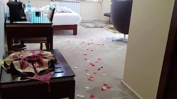 Habitación de hotel con cama, escritorio, silla y sofá. Pétalos de rosa esparcidos por el suelo.