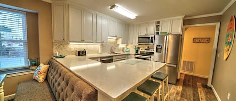 A modern kitchen with white cabinets, stainless steel appliances, a large island with bar stools, and a view of the street through a window.
