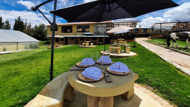 An outdoor dining area with stone tables and benches, surrounded by a well-maintained lawn and a building with a tiled roof. - CLUB EL LLANO (Cómbita)