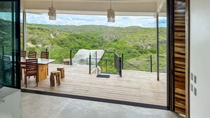 Un balcón espacioso con una mesa y sillas de madera, una roca grande y vistas a colinas verdes.