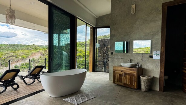 Un baño moderno con una bañera independiente, un tocador de madera y un ventanal con vistas a un paisaje montañoso.