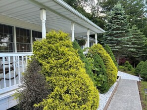 Enjoy a serene outdoor space with a covered porch surrounded by lush evergreen shrubs and a majestic coniferous tree.