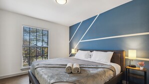 Una habitació d'hotel moderna amb un llit gran, una finestra amb persianes, una tauleta de nit amb una làmpada i una paret d'accent blava.