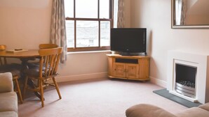 Un salon avec un téléviseur posé sur un meuble en bois, une cheminée, une table à manger avec des chaises et une fenêtre avec des rideaux.