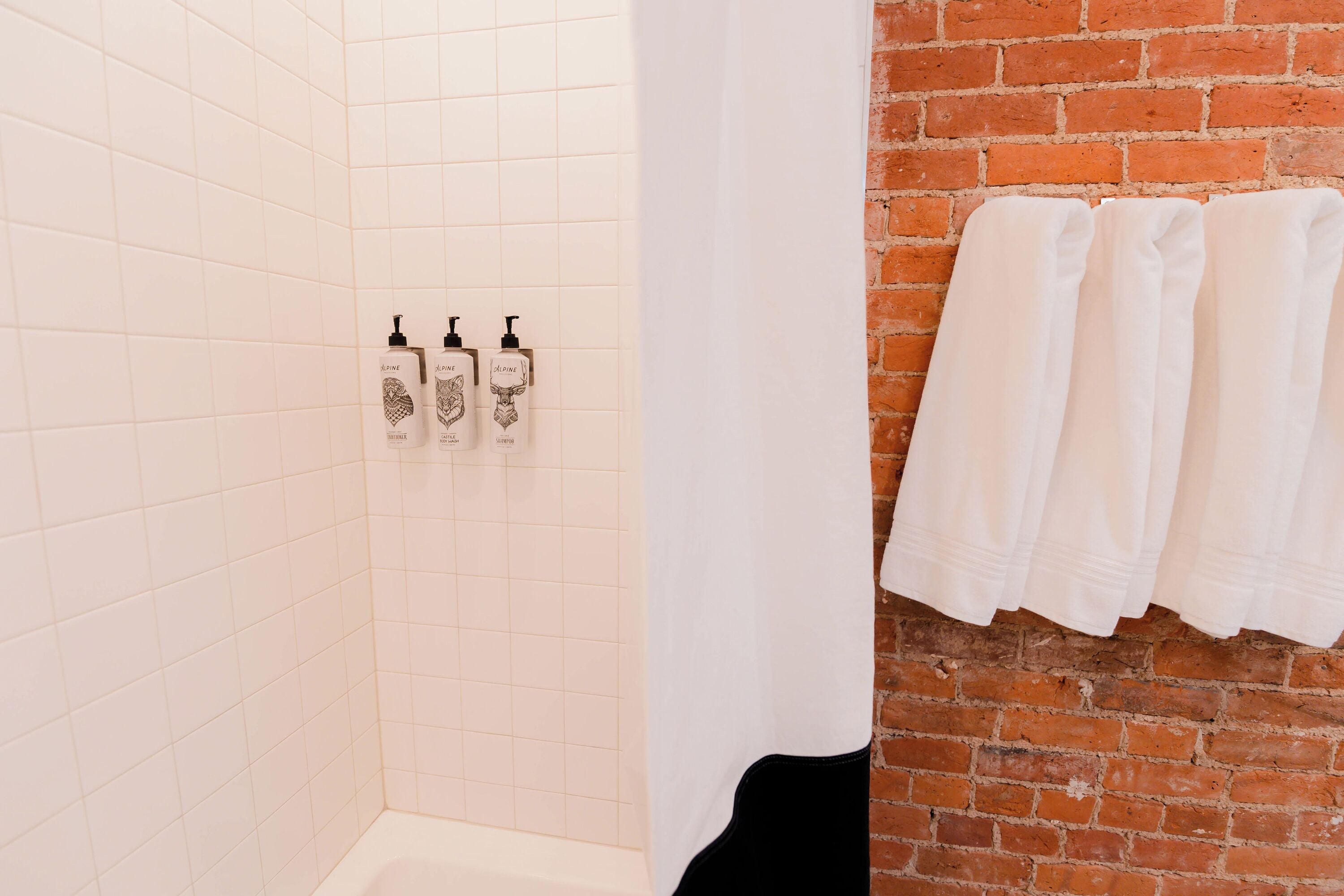 Un baño con paredes de azulejos blancos, una pared de ladrillo y tres botellas de jabón para manos en la pared.