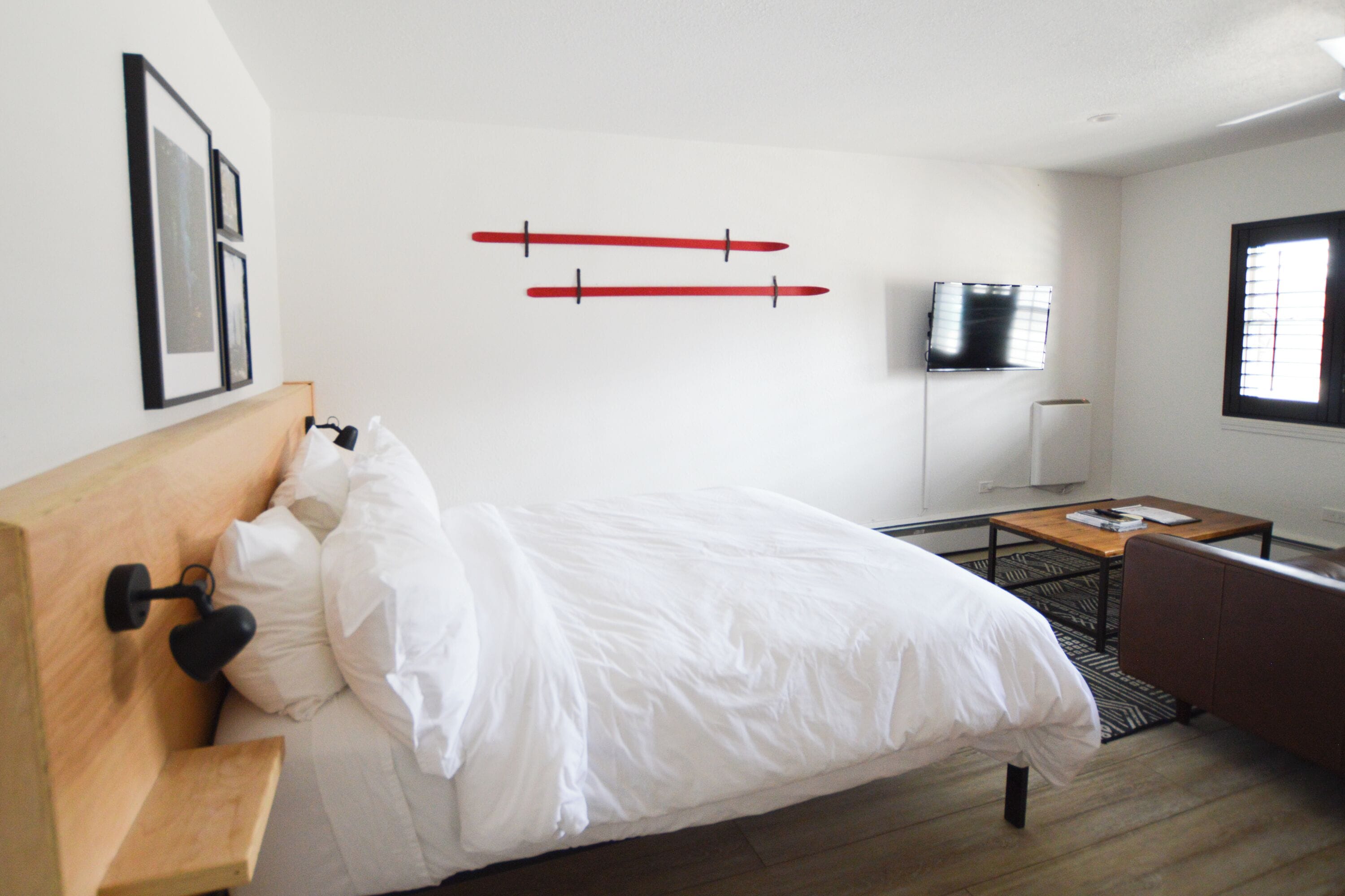 Una camera d'albergo con un letto, una testiera in legno, un piccolo tavolo, una poltrona e una TV a parete, oltre a una finestra con persiane.