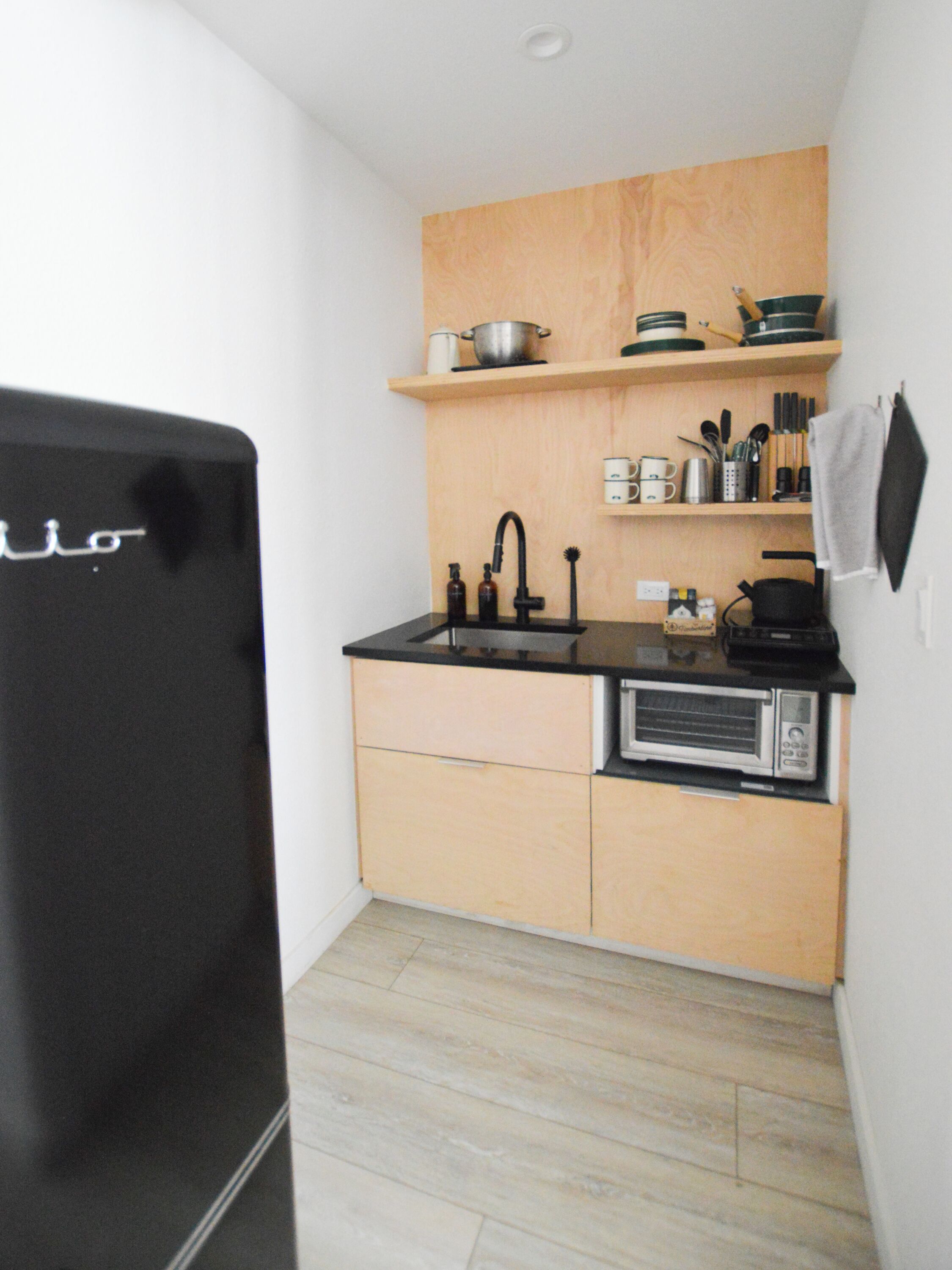 A compact kitchen with a black refrigerator, wooden cabinets, a black countertop, and a microwave.