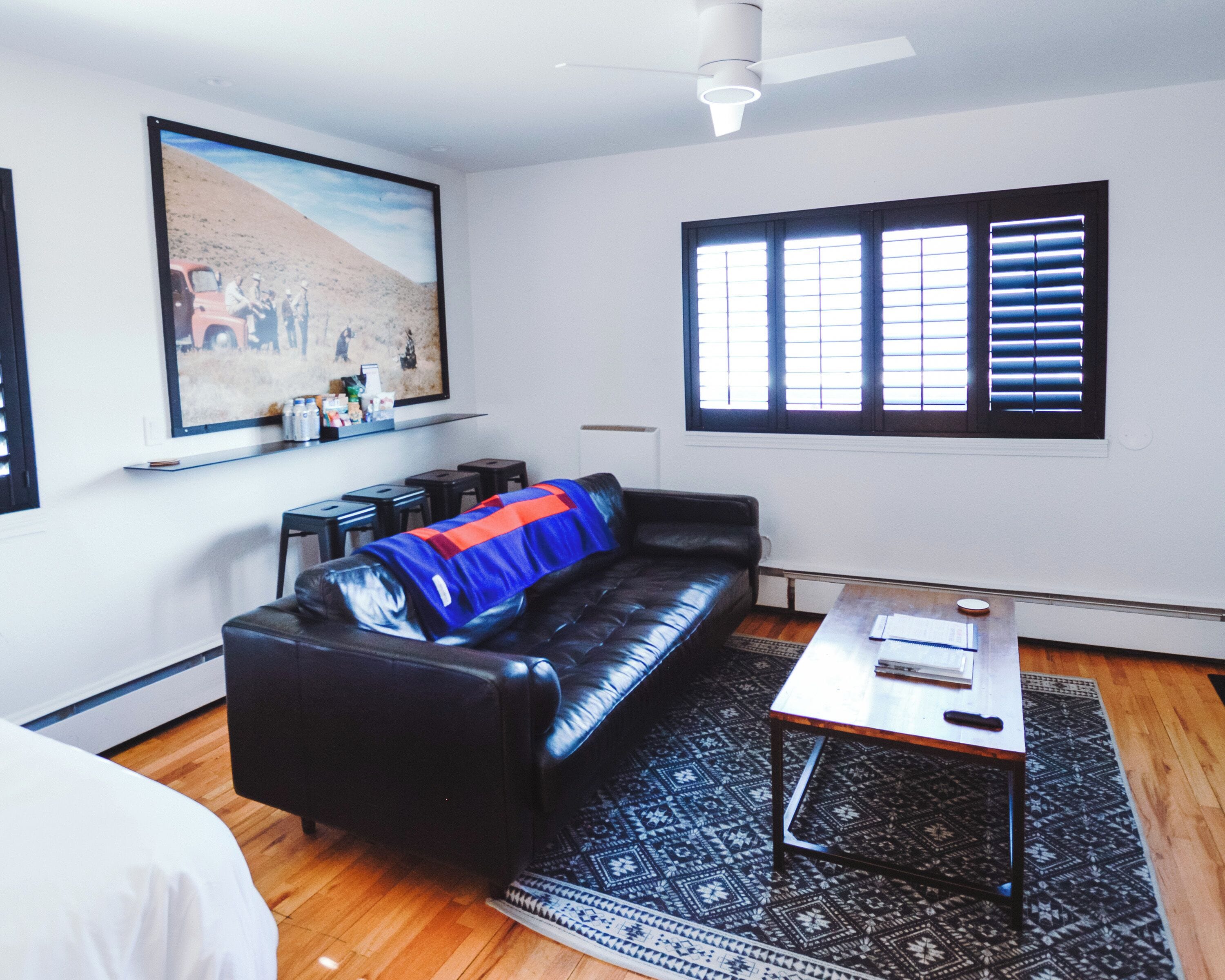 A living room with a black leather sofa, a patterned rug, a coffee table, a painting on the wall, and a window with blinds.