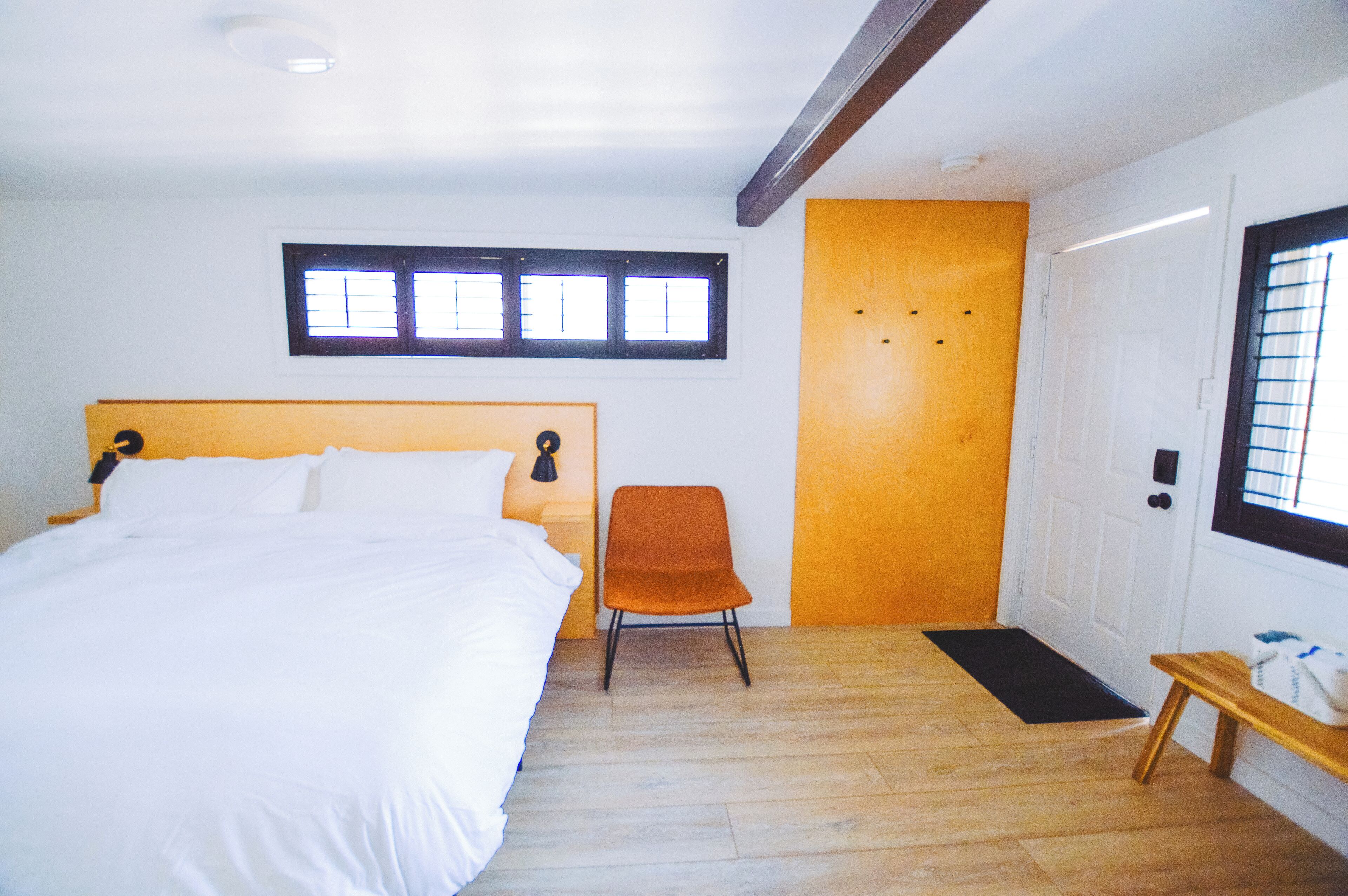 Une chambre à coucher avec un lit, une chaise orange, un banc en bois, une porte et une fenêtre avec des stores.