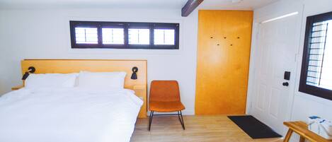 Une chambre à coucher avec un lit, une chaise orange, un banc en bois, une porte et une fenêtre avec des stores.