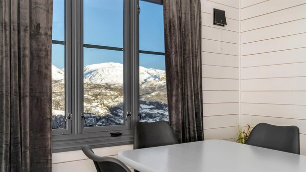 Habitación con un ventanal grande que ofrece vistas a montañas nevadas. Hay una mesa con sillas y una pequeña planta en el alféizar.