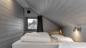 Una habitación pequeña con techo inclinado, dos camas, un ventanal con cortina y una lámpara fijada a la pared.