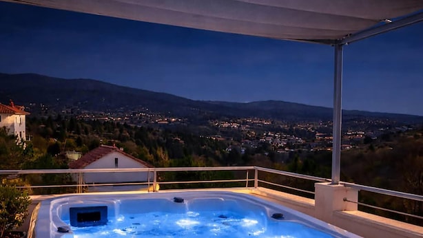A hot tub on a balcony with a view of a town and mountains at dusk.